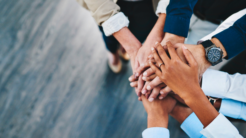 Photo displays a group of hands on top of each other. A team huddle.